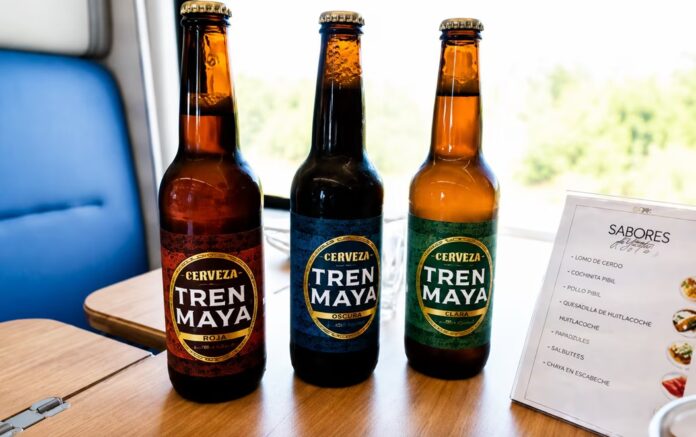 Tres botellas de Cerveza Artesanal Tren Maya exhibidas sobre una mesa dentro de un vagón ferroviario, producto elaborado por una empresa privada.