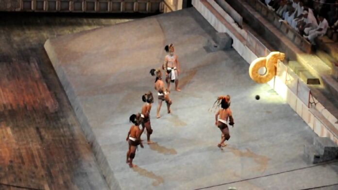 Jugadores participan en una representación del ancestral juego maya Pok Ta Pok dentro de una cancha ceremonial con aro de piedra iluminado.