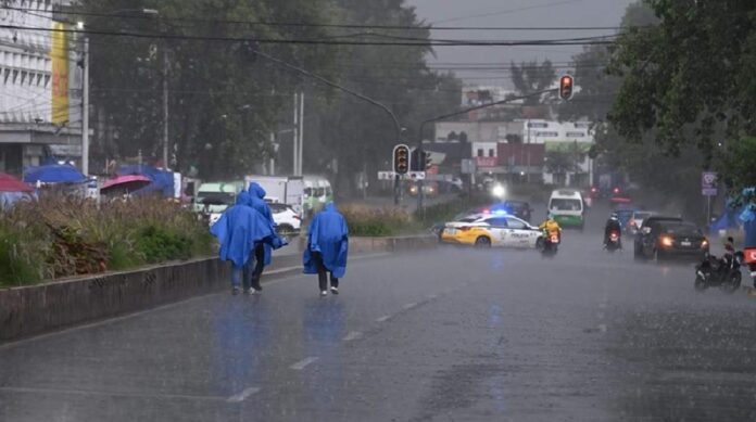 Personas caminan bajo una intensa lluvia en una avenida con encharcamientos, mientras vehículos circulan en condiciones de baja visibilidad durante una tormenta.
