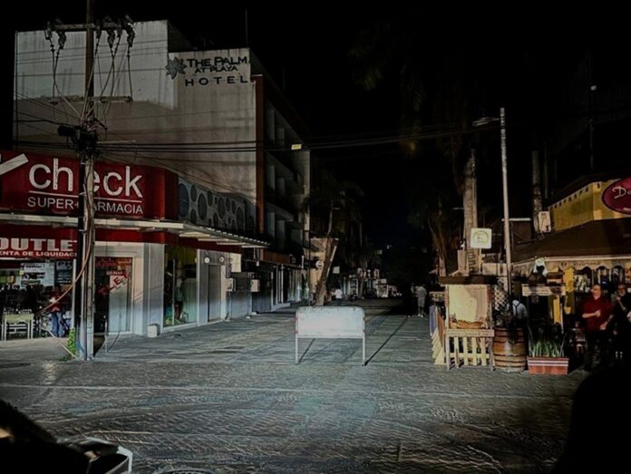 Calle del centro de Playa del Carmen durante la noche sin alumbrado público, con comercios, un hotel y barricadas visibles.