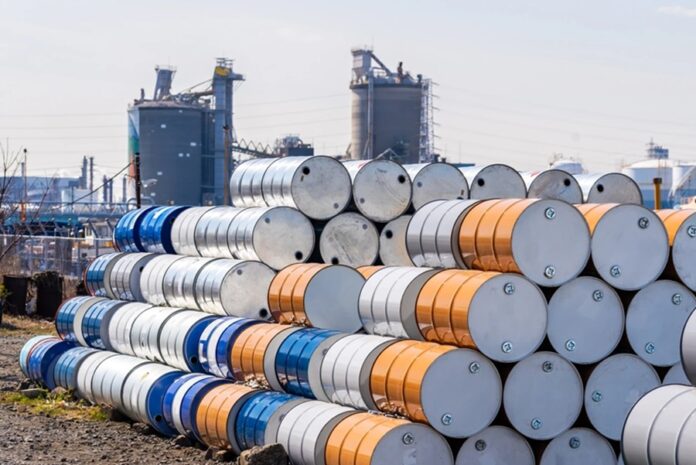 Barriles metálicos apilados frente a instalaciones industriales relacionadas con la industria petrolera.