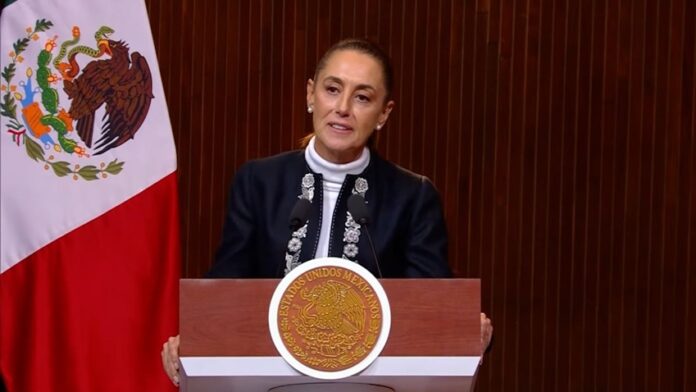 La presidenta Claudia Sheinbaum ofrece un mensaje desde un podio con el escudo nacional y la bandera de México.
