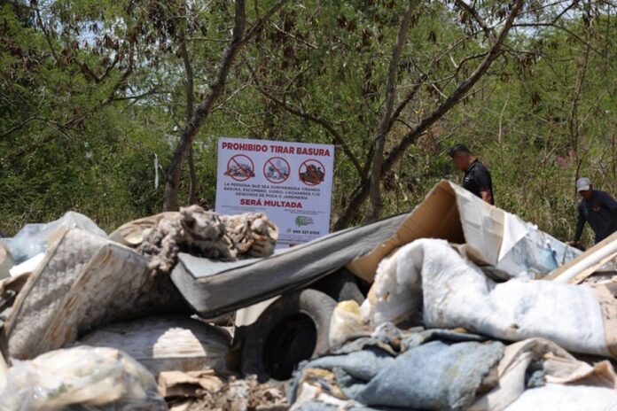 Montones de basura acumulada en un basurero clandestino de la Supermanzana 200 de Cancún, con un letrero que prohíbe tirar residuos y personal municipal al fondo.