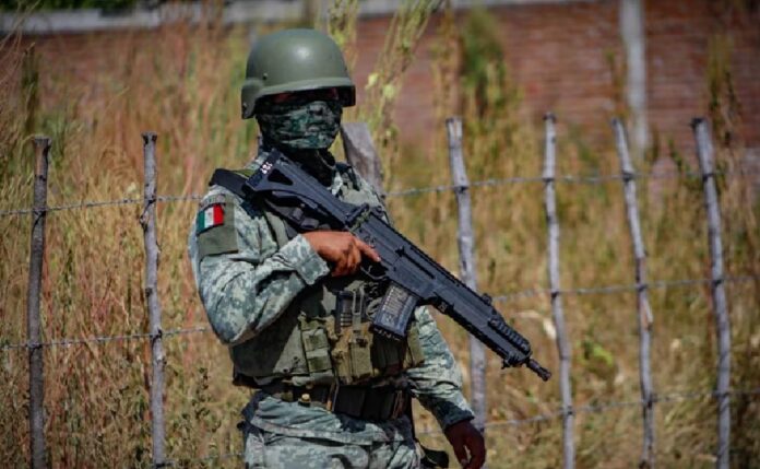 Elemento del Ejército Mexicano con uniforme, casco y arma larga permanece de pie junto a una cerca de alambre.