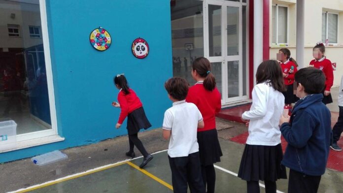 Estudiantes participan en una actividad al aire libre dentro del patio de una escuela mientras forman una fila frente a una pared azul.