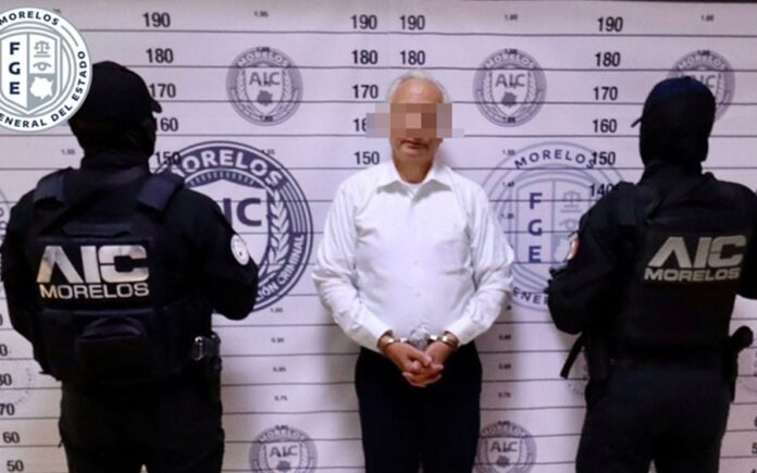 Víctor Rodríguez Padilla permanece esposado frente a un muro de identificación, custodiado por dos agentes de la Agencia de Investigación Criminal de Morelos.