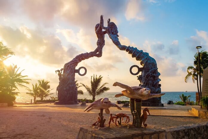 Escultura del Portal Maya frente al mar en Playa del Carmen durante el atardecer, con palmeras y cielo parcialmente nublado. Foto: Gobierno de Playa del Carmen.