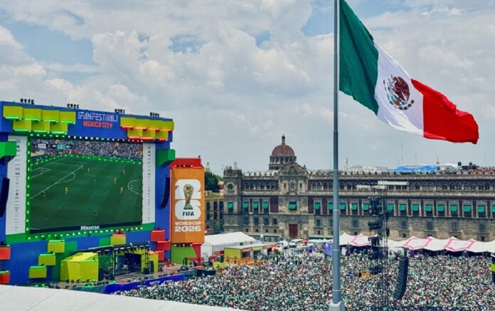 Pantalla gigante del Fan Fest en el Zócalo de la Ciudad de México con aficionados reunidos y una bandera monumental de México.