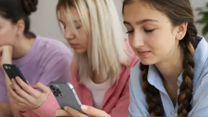 Tres adolescentes observan y utilizan teléfonos celulares mientras permanecen sentadas en un salón.