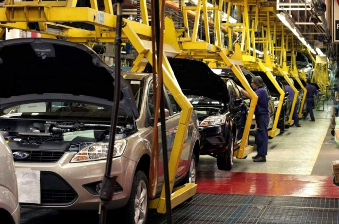 Vehículos Ford avanzan por una línea de ensamblaje mientras trabajadores realizan labores dentro de una planta de producción automotriz.