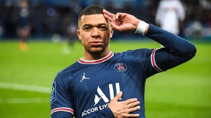 Kylian Mbappé con el uniforme de Francia realiza un saludo con la mano mientras permanece sobre la cancha.