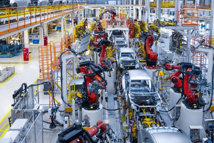 Línea automatizada de ensamblaje de vehículos en una planta industrial con brazos robóticos y carrocerías en producción.