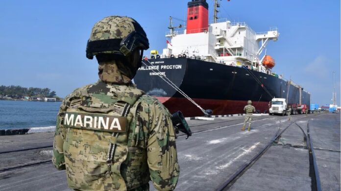 Elementos de la Secretaría de Marina resguardan un buque tanque atracado en un puerto, mientras vehículos de carga permanecen en el muelle.