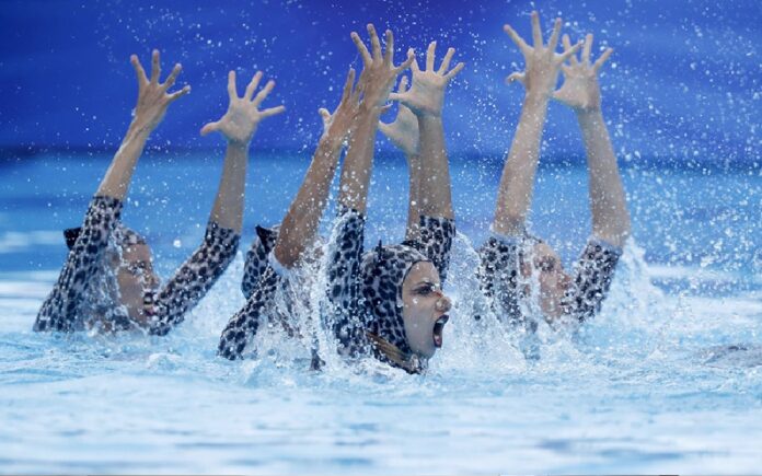 Nadadoras de natación artística realizan una rutina dentro de una piscina mientras levantan los brazos entre salpicaduras de agua.