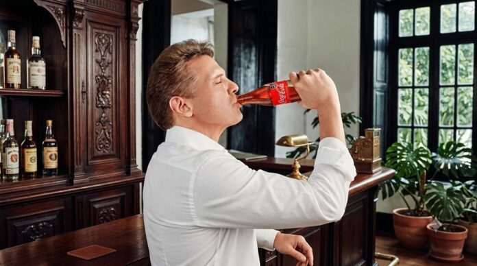 Luis Miguel sostiene una botella de refresco mientras bebe durante la grabación de un video promocional en un salón con mobiliario de madera.